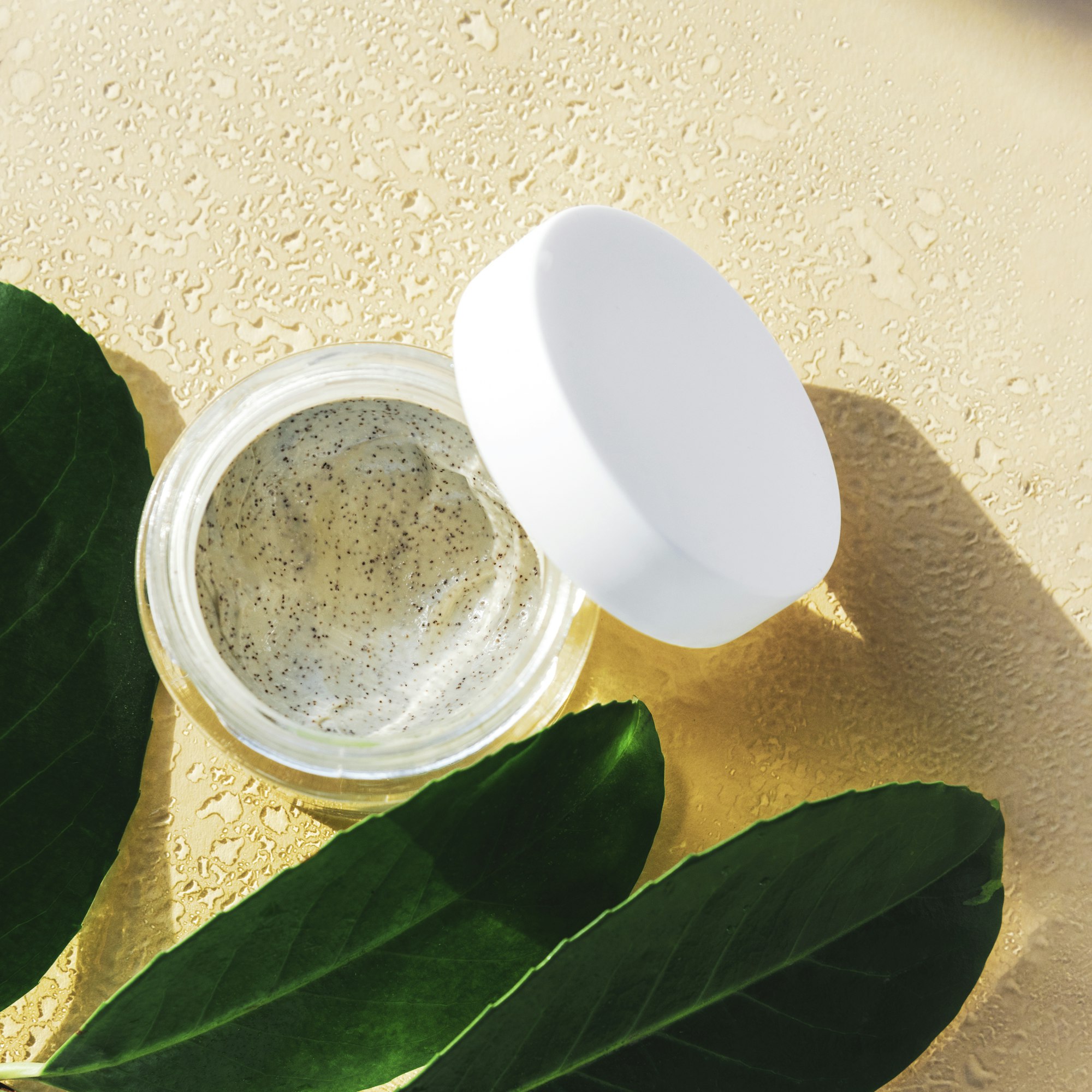 Face peeling mask in a jar and green leaves, neutral background with water drops.